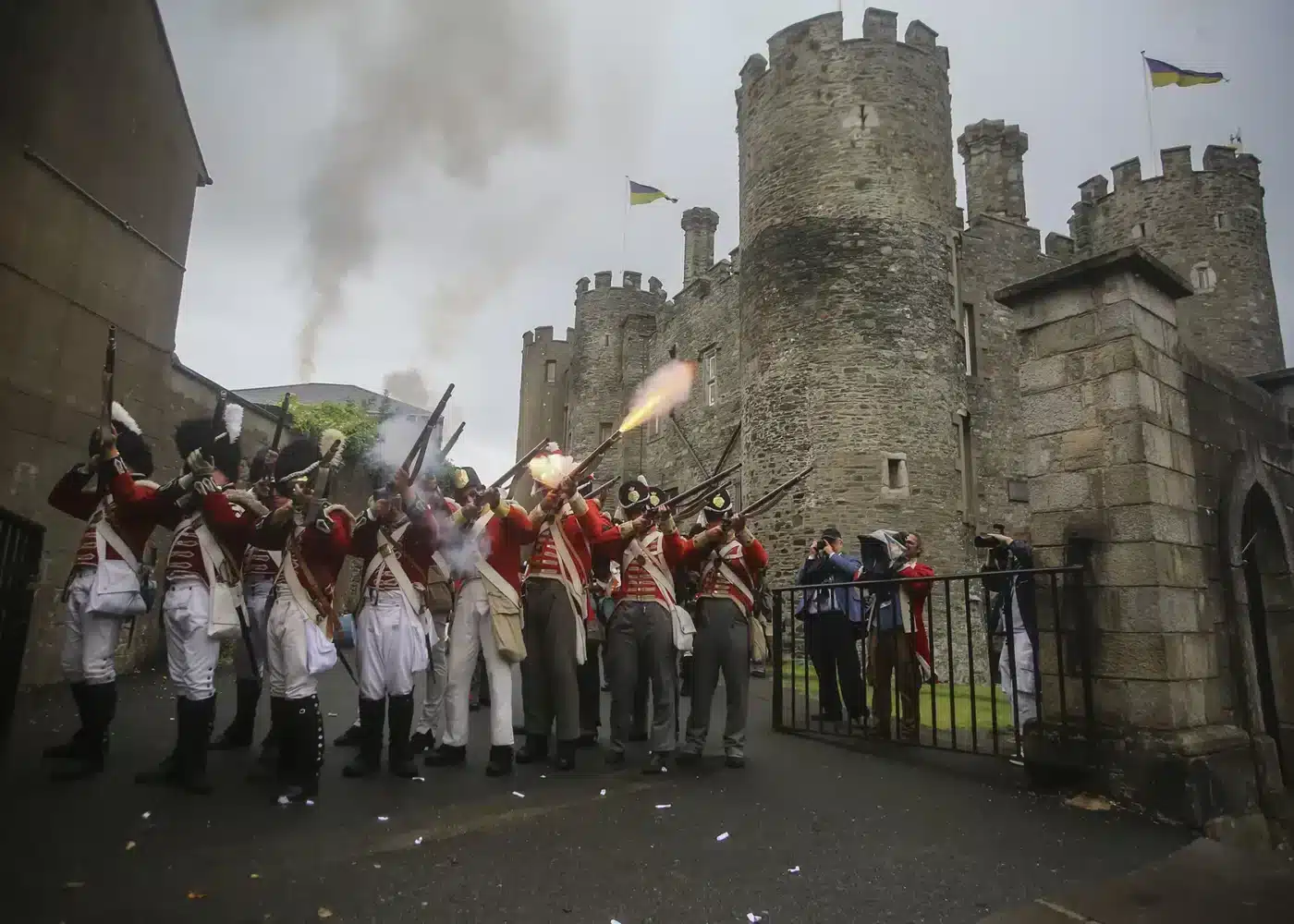 Historical reenactors in red military uniforms firing muskets outside the stone towers of Enniscorthy Castle during a battle demonstration, with smoke rising around them.