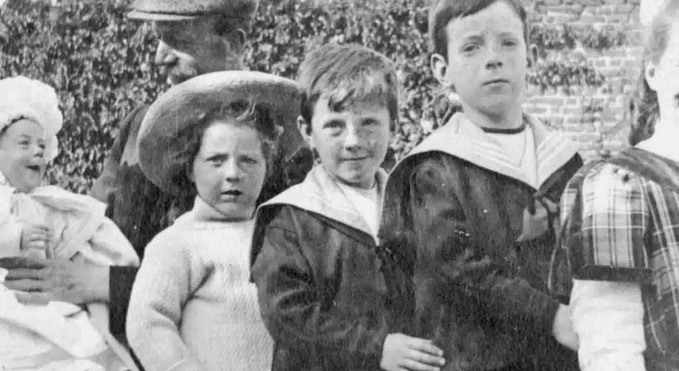 Vintage black and white photo of four young children standing in a row, with an adult holding a baby on the left, all dressed in early 20th-century clothing.