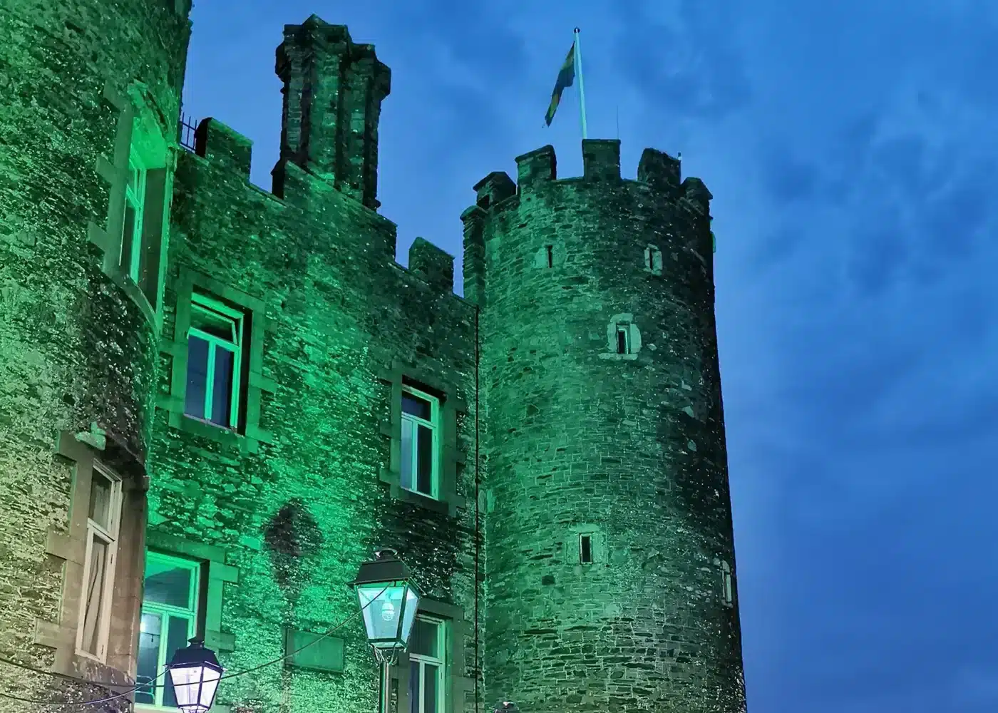 Night-time view of Enniscorthy Castle illuminated in green, showing its stone tower, windows and Irish flag against a deep blue sky.