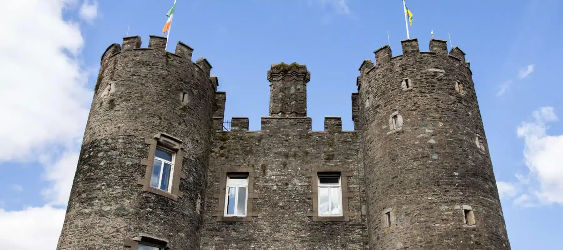 Front view of Enniscorthy Castle’s stone towers with flags flying against a blue sky with white clouds.