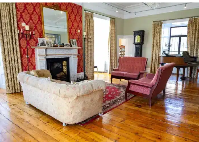 Elegant period room in Enniscorthy Castle featuring vintage sofas, a marble fireplace with a large mirror, wooden floors, tall, draped windows and a piano in the corner.