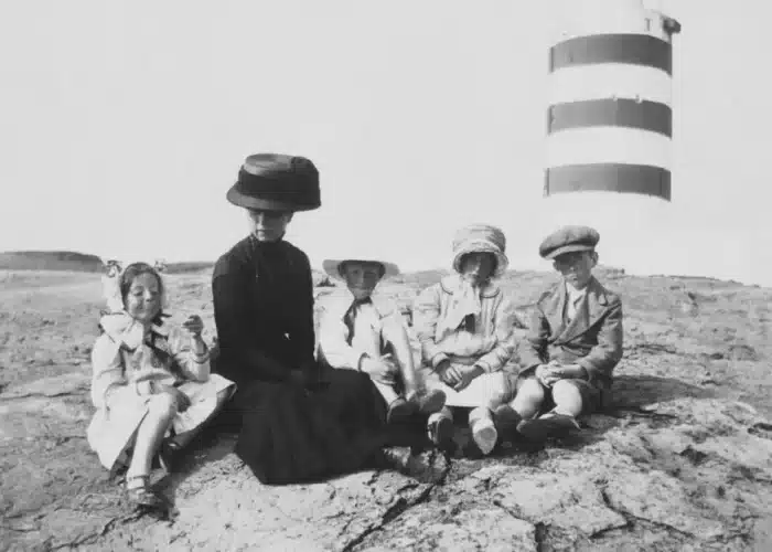 Historic black and white photograph of Josephine Roche and four children sitting on rocky ground with a lighthouse in the background.