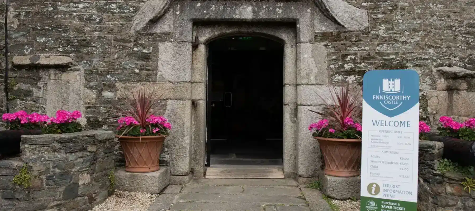 Entrance to Enniscorthy Castle with stone doorway, potted pink flowers, and a welcome sign beside the path.