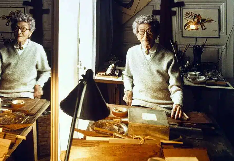 An older Eileen Gray standing in a studio-style workspace surrounded by design materials, with her reflection visible in a large mirror.