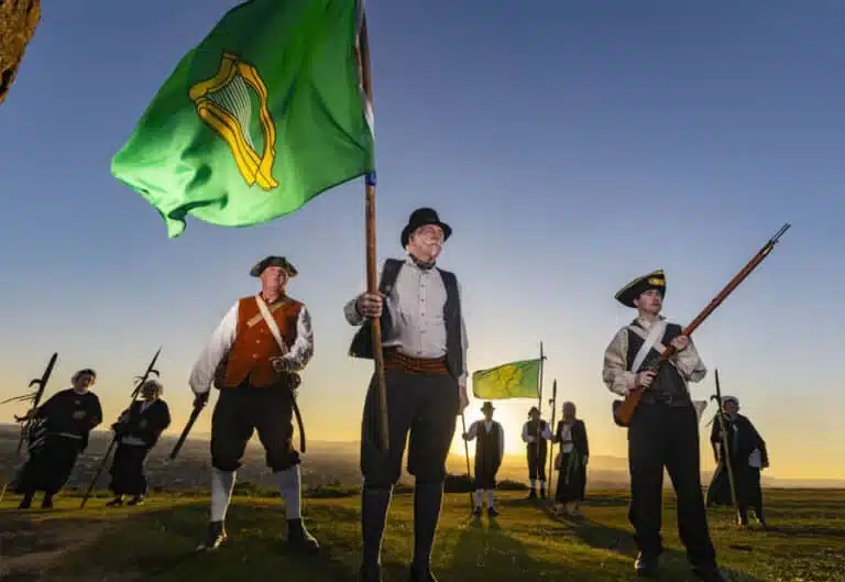 Reenactors dressed as 1798 Irish rebels standing on a hillside at sunset, holding period weapons. And green flags with a gold harp.