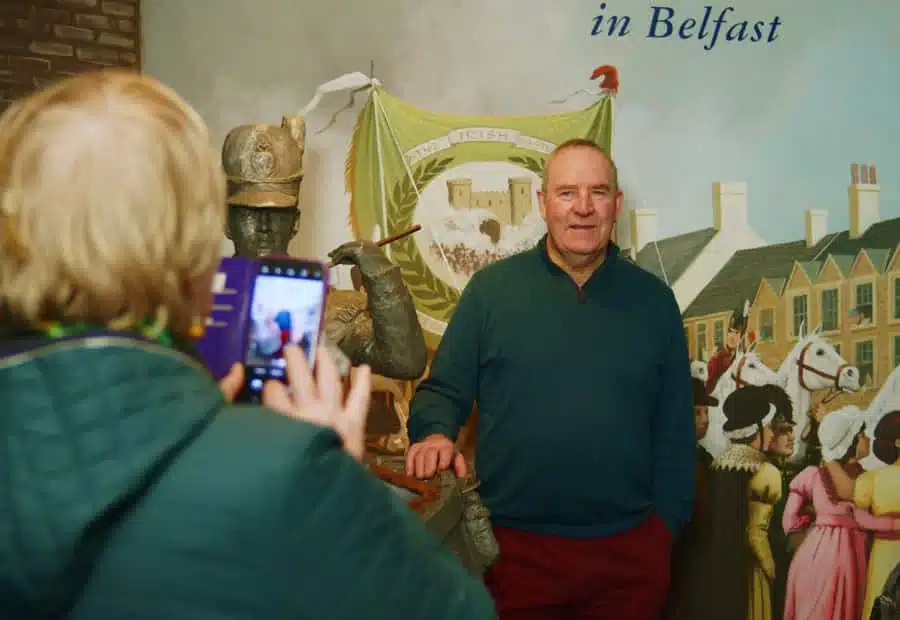 Person taking photo on phone in the National 1798 Rebellion Centre, Enniscorthy, of a man standing in front of a historical mural and exhibit which features period banners and artefacts.