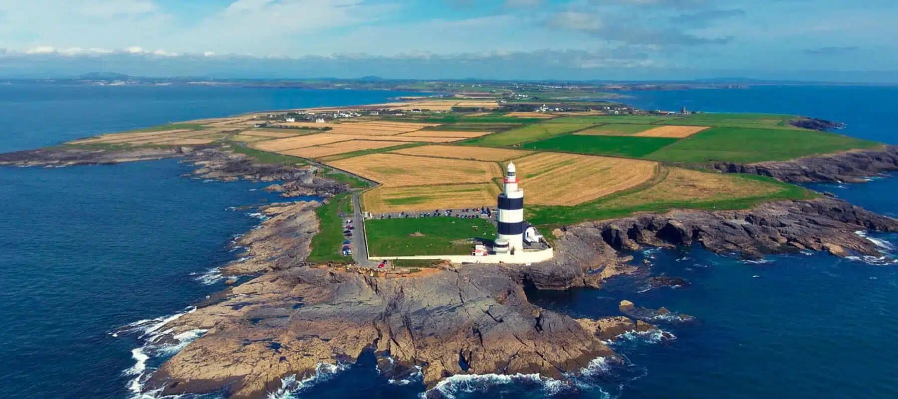 Aerial view of Hook Lighthouse on a rocky peninsula, surrounded by fields and the Atlantic Ocean.